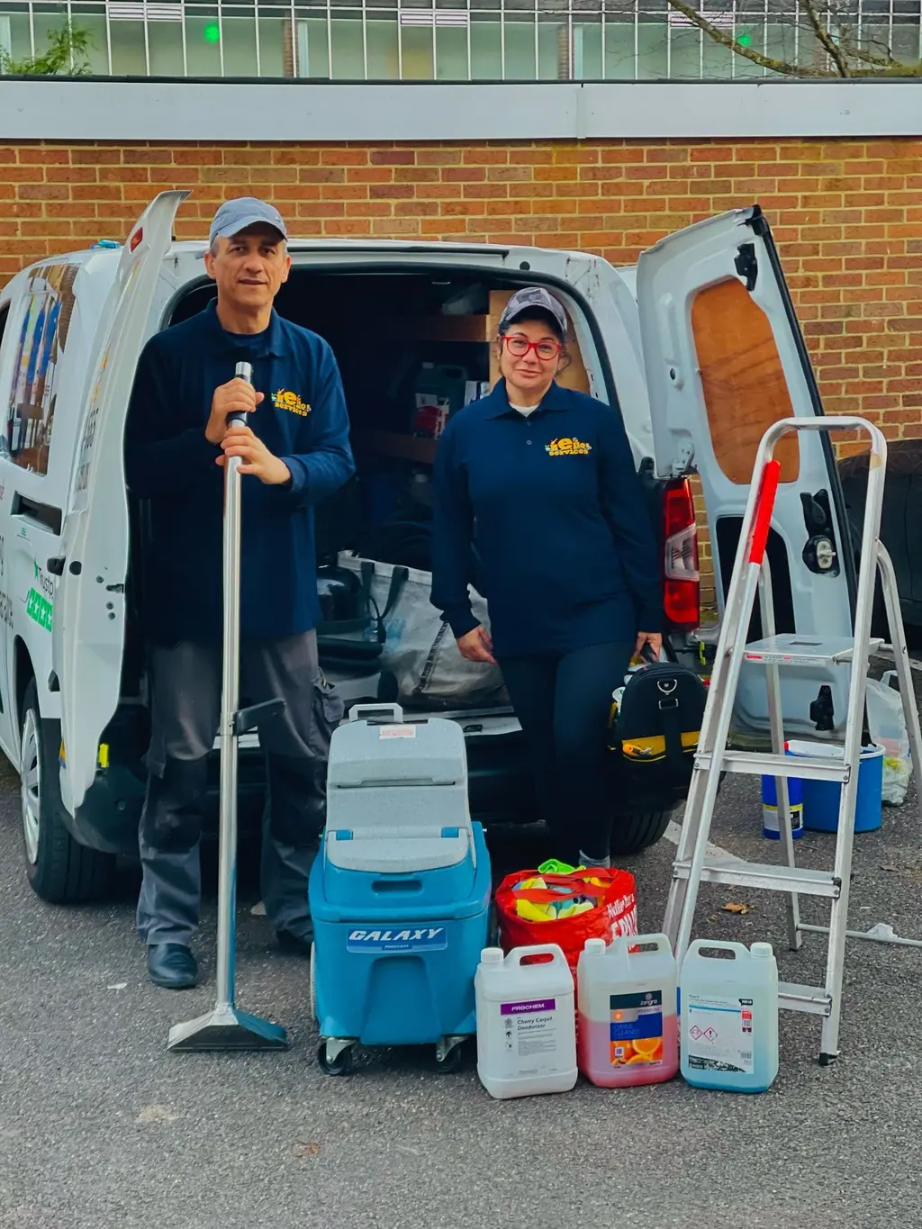 Two Hello Services cleaners in navy uniforms beside their open van with a carpet extraction machine, cleaning solutions and a stepladder